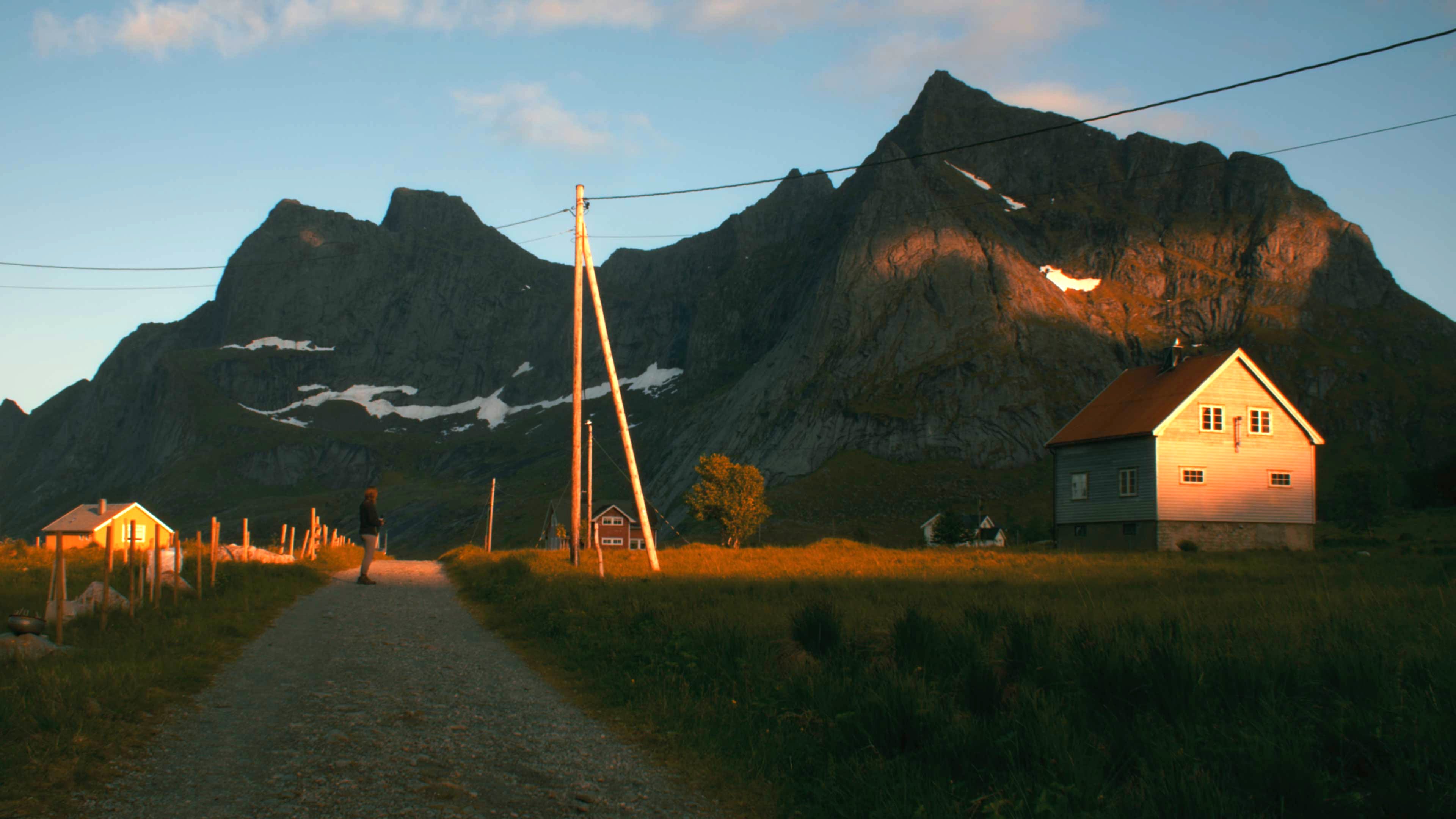 A gravel path leads through a coastal settlement at golden hour, timber houses lit warm beneath a dark granite peak