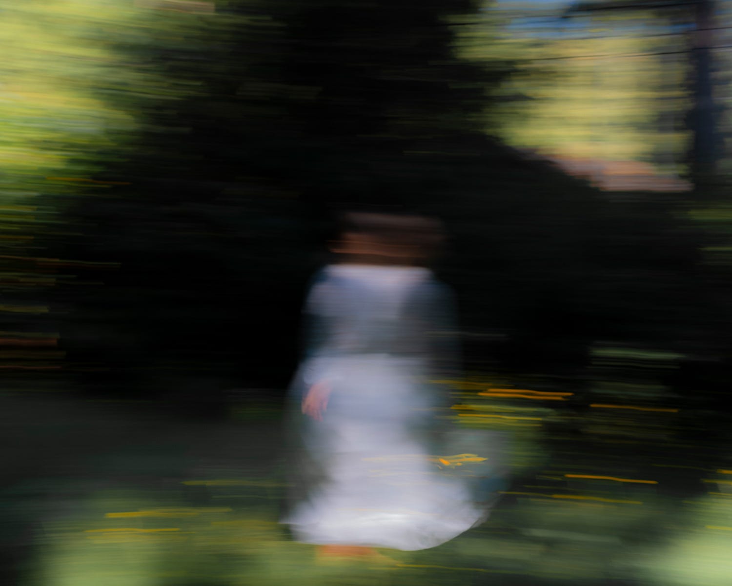 A figure in a pale dress, blurred by long exposure, moving through green shade toward dark trees